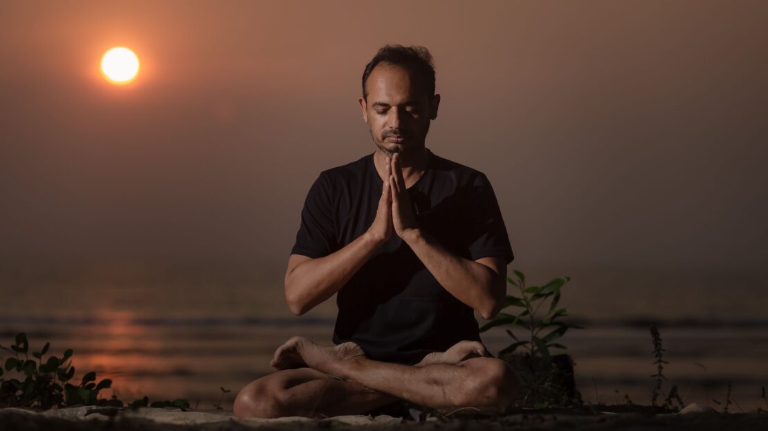 Man seated in Padmasana at sunrise with hands in prayer, meditating by the ocean to honour the roots of yoga on International Yoga Day