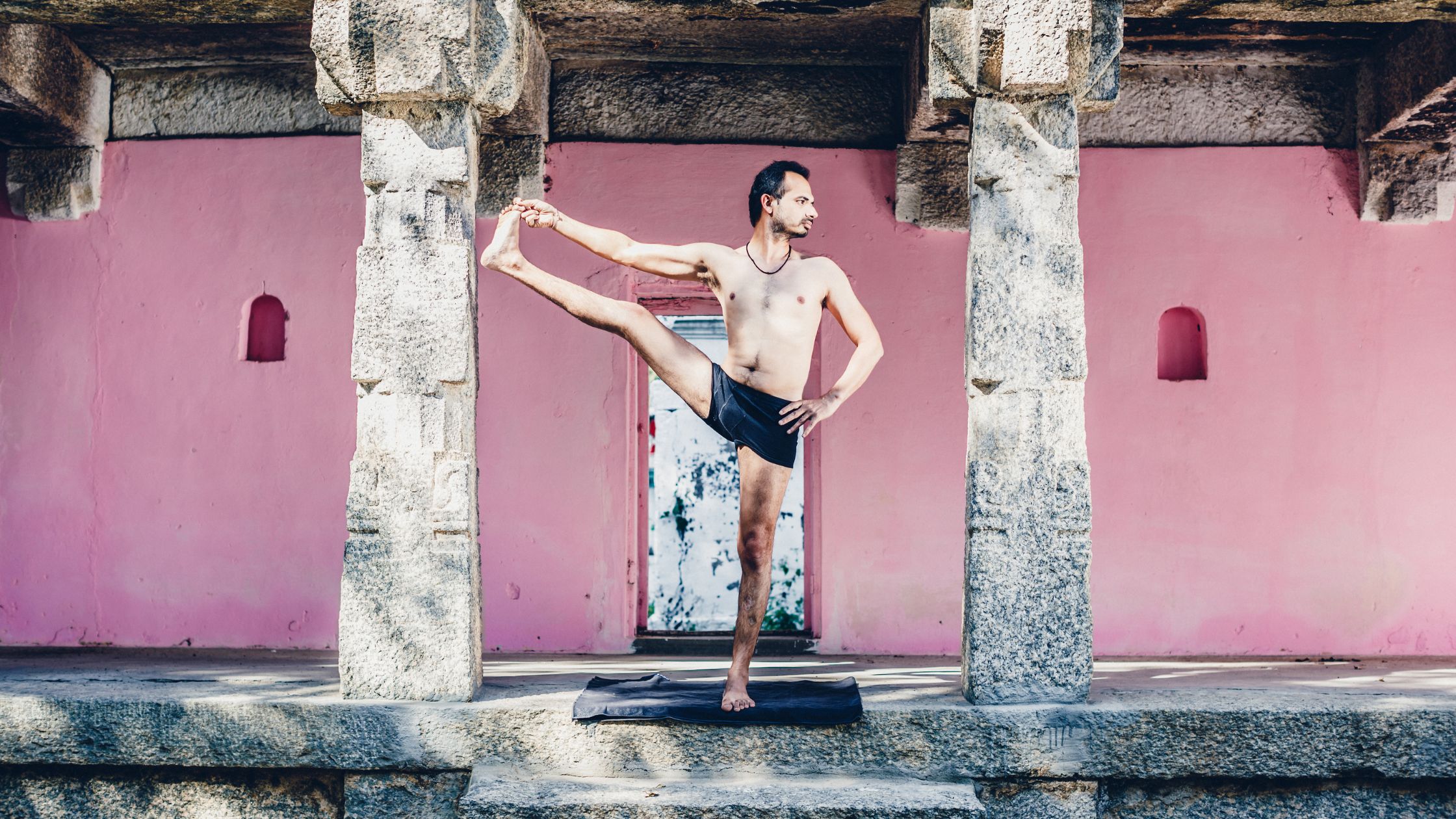 Yogacharya Lalit practising an extended leg stretch in front of a pink temple wall, representing lineage and mastery
